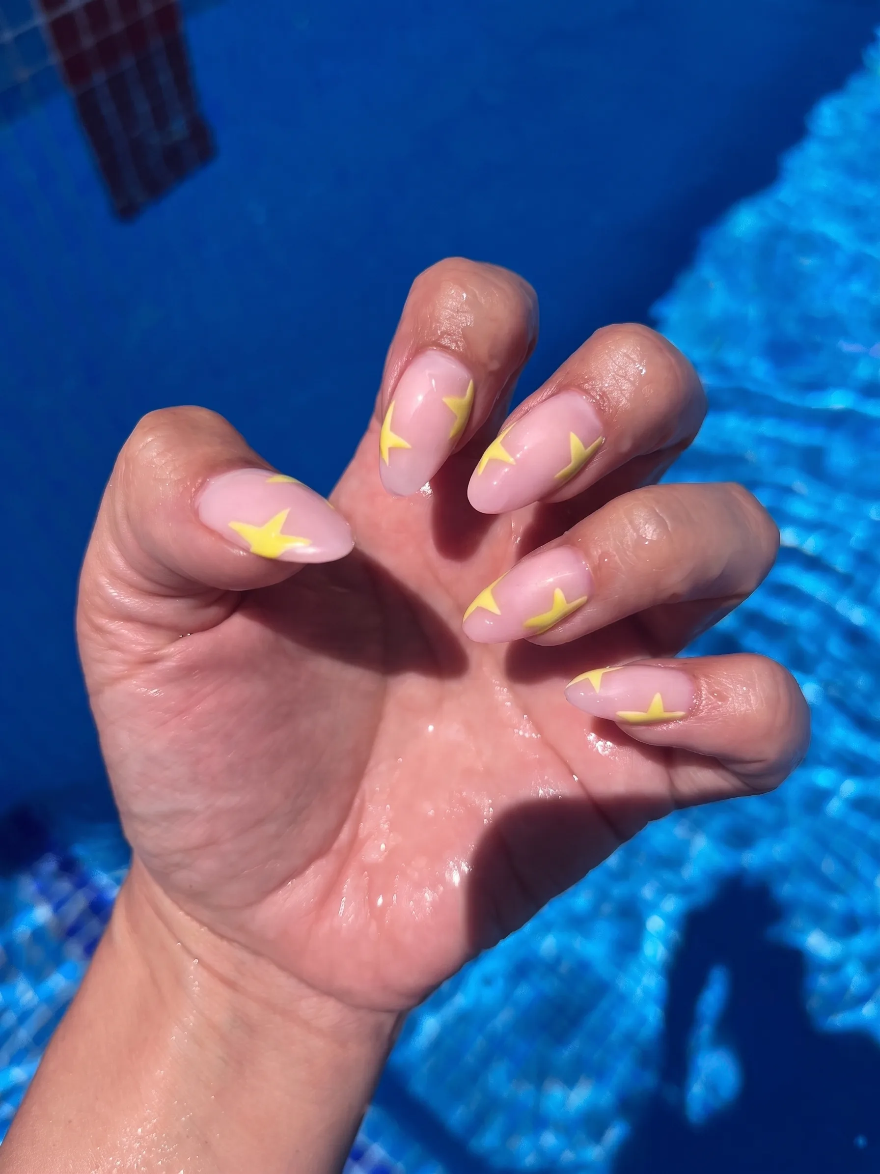Transparante nude amandelnagels met botergele steraccenten, gefotografeerd bij het zwembad.