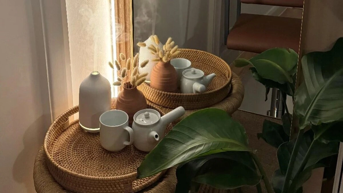 Close-up van een rieten dienblad met een diffuser en een theeservies naast een grote groene plant in een salon