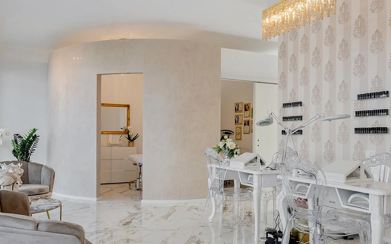 Het elegante interieur van MC Beauty Center met een vloer in marmerlook, witte gebogen wanden en een gouden kristallen kroonluchter. Rechts staat een nagelstation met ghost chairs, terwijl fluwelen fauteuils en een goudomlijste doorgang naar een behandelkamer leiden.