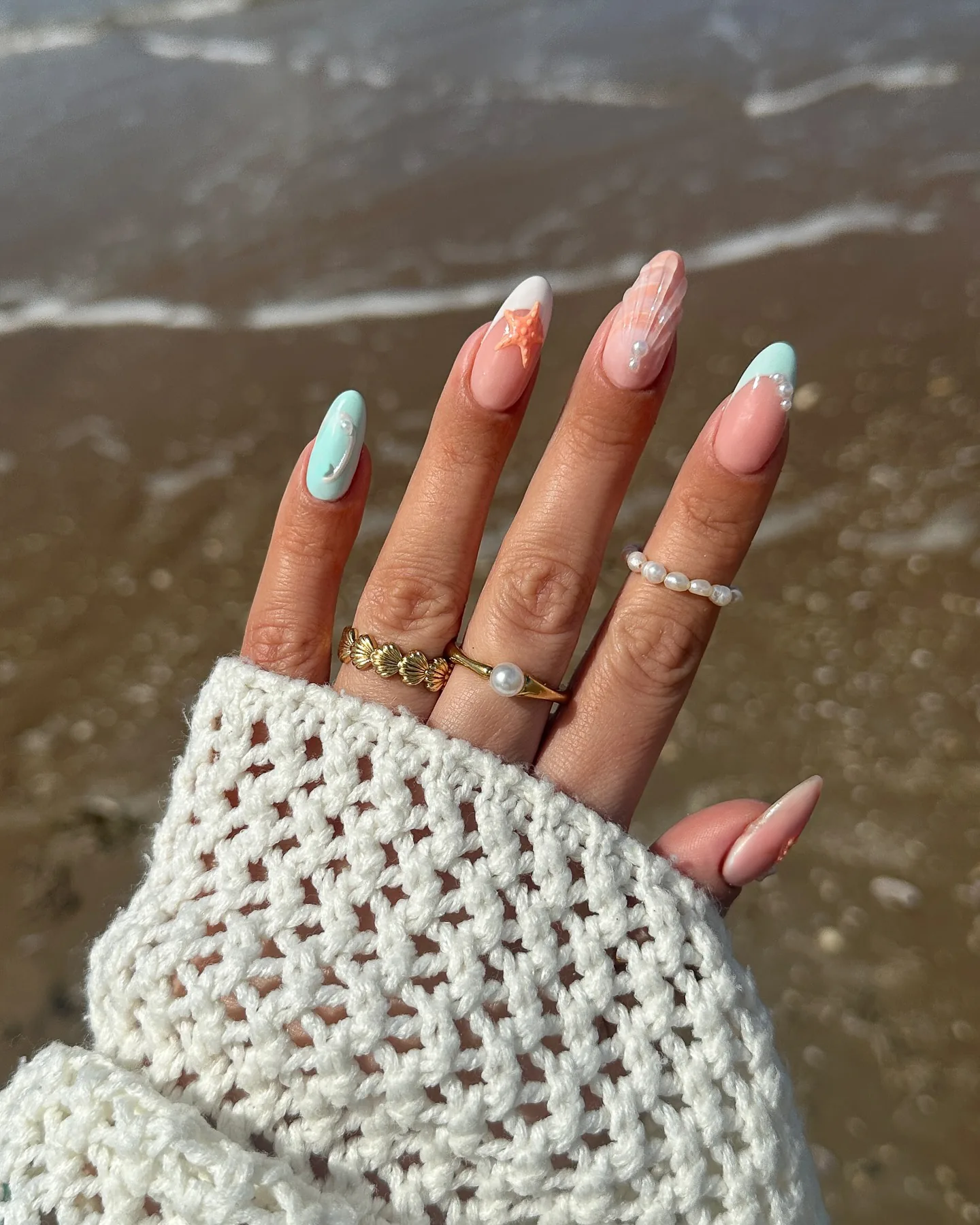 Amandelnagels met pastelkleurige mermaid-art – een zeester, parelaccenten en een parelmoerglans – tegen nat zand.