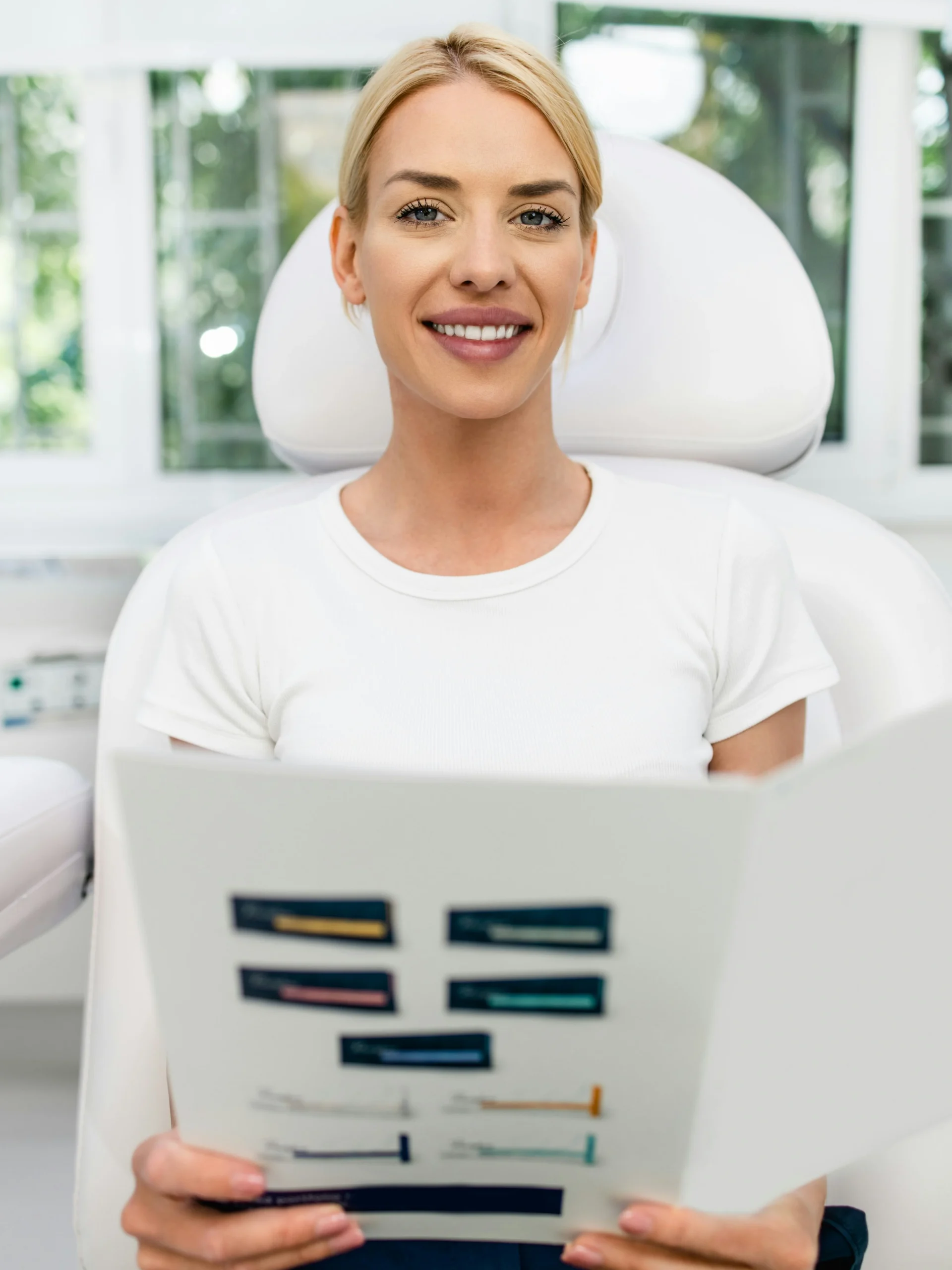 Een cliënt zit in een lichte, schone kliniek en glimlacht terwijl ze een informatiebrochure over gezichtsbehandelingen vasthoudt