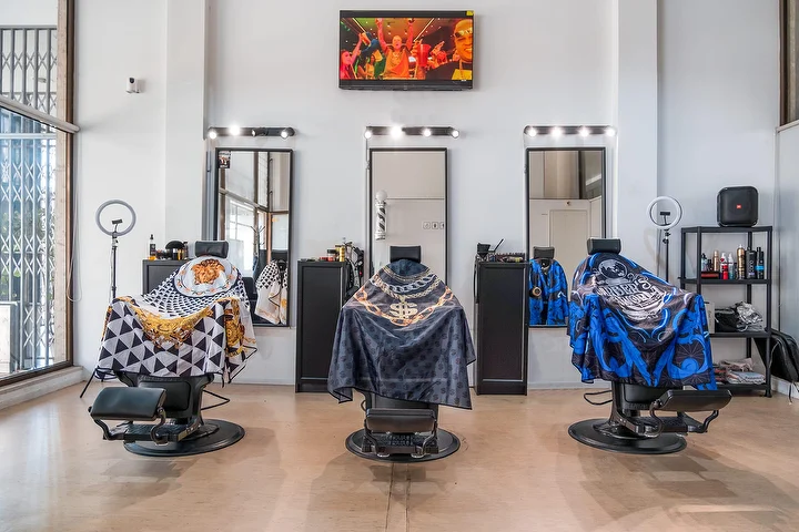 Een lichte barbershop met hoge plafonds en drie zwarte barbiersstoelen met gedessineerde capes, elk voor een spiegel omlijst door lampjes, met ringlampen, een wandscherm en hoge ramen.