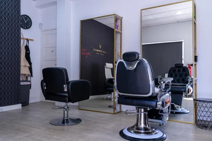 Een sober ingericht saloninterieur met een zwarte stylingstoel en een barbiersstoel van chroom en leer op een lichte houten vloer, een grote spiegel met gouden lijst, een wandklok en schorten naast een donkere accentmuur.