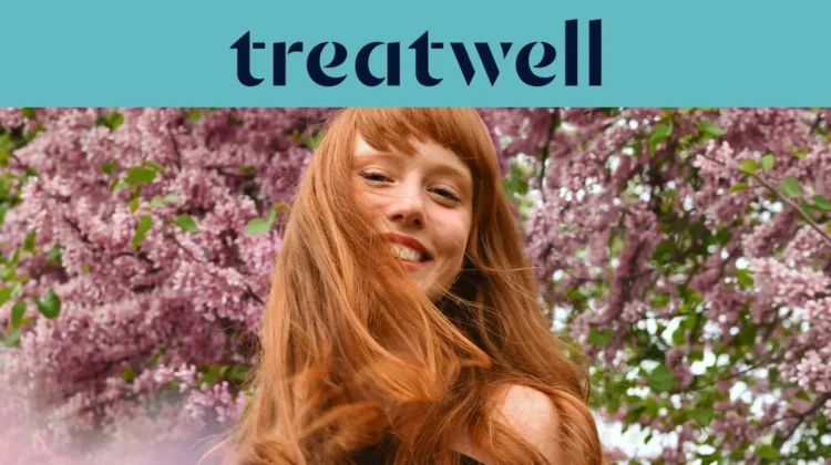 Close-up portret van een glimlachende jonge roodharige vrouw met een pony, wiens haar in de wind waait voor een achtergrond van roze seringentakken. Een lichtblauwe banner bovenaan toont het "Treatwell"-logo in het donkerblauw.