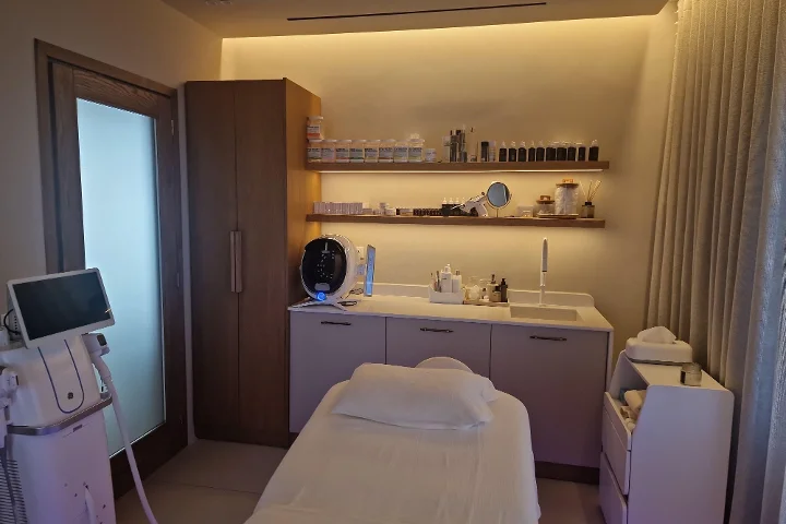 Een warm verlichte behandelkamer met een witte gezichtsbehandeltafel en netjes opgevouwen handdoeken, donkere houten schappen vol huidverzorgingsproducten, een huidanalysescherm en behandelapparatuur ernaast, klinisch maar gezellig.