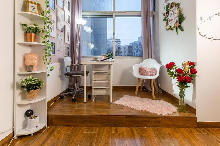 Een kleine schoonheidsstudio op een verhoogd houten platform, met een wit bureau en een klemlamp, een bureaustoel en een fauteuil, hoekplanken met planten, een vaas met rode rozen en een raam op de stad.