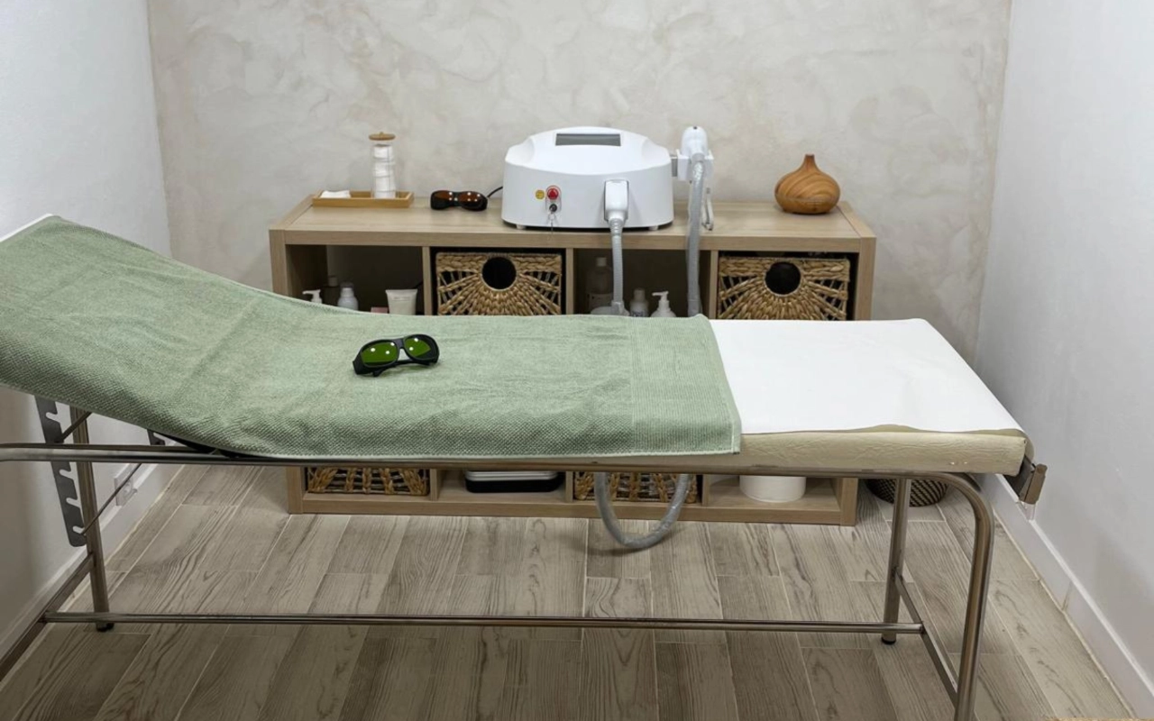 Een neutraal getinte behandelkamer met een metalen bed met een saliegroene handdoek, een donkere beschermbril erbovenop, een laserapparaat op een houten console en gevlochten opbergmanden eronder.