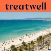 Een luchtfoto van Playa de Palma op Mallorca, met een lange boog van goudkleurig zand, rijen parasols, kalm turquoise water en een met palmen omzoomde boulevard langs de stad, met heuvels in de verte onder een strakblauwe lucht.