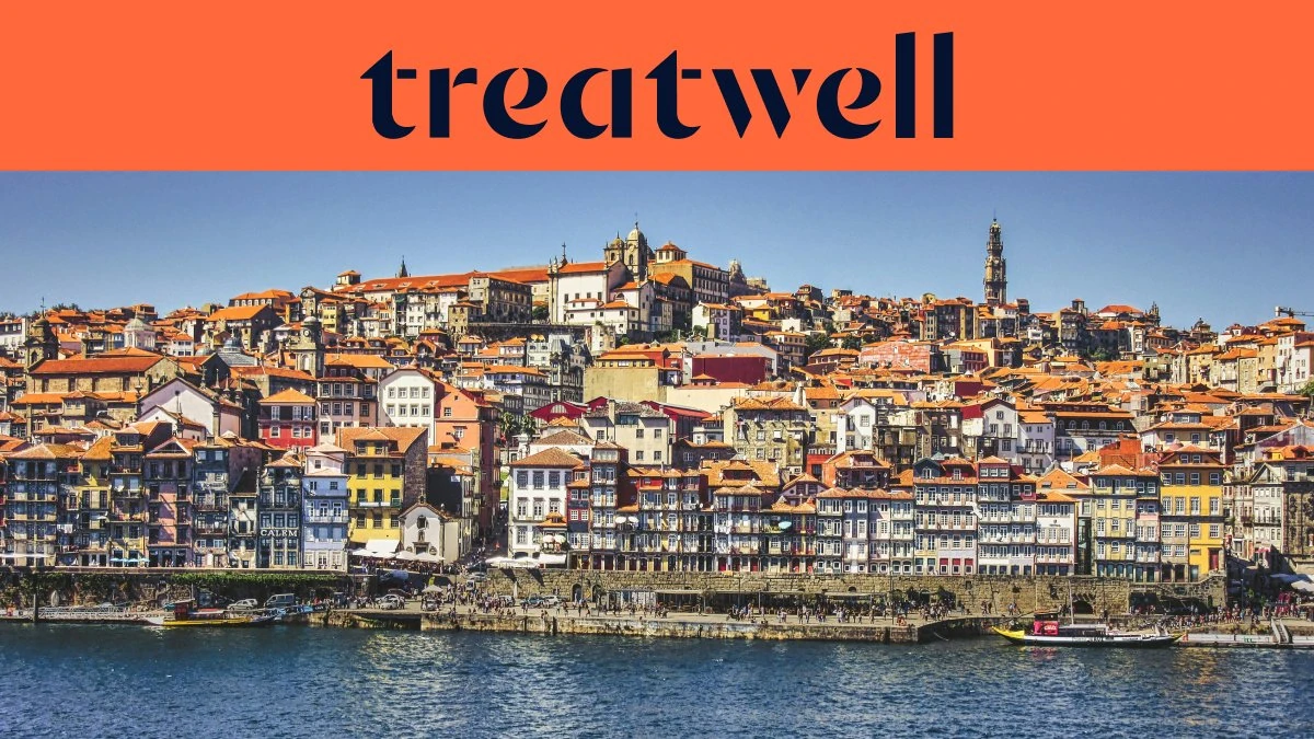 De Ribeira-kade in Porto, met rijen kleurrijke huizen en terracotta daken die vanaf de rivier de Douro tegen de heuvel op lopen, en de Clérigos-toren aan de horizon onder een strakblauwe lucht.