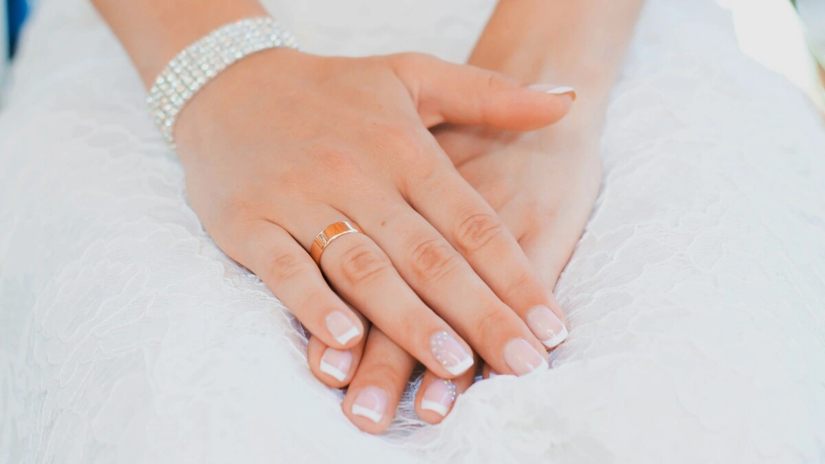 De handen van een vrouw rusten in haar schoot; ze draagt een kanten trouwjurk en een verzorgde French manicure in een klassieke combinatie van nude en wit op korte, vierkant-ovale nagels, met een fijne versiering op de ringvingers