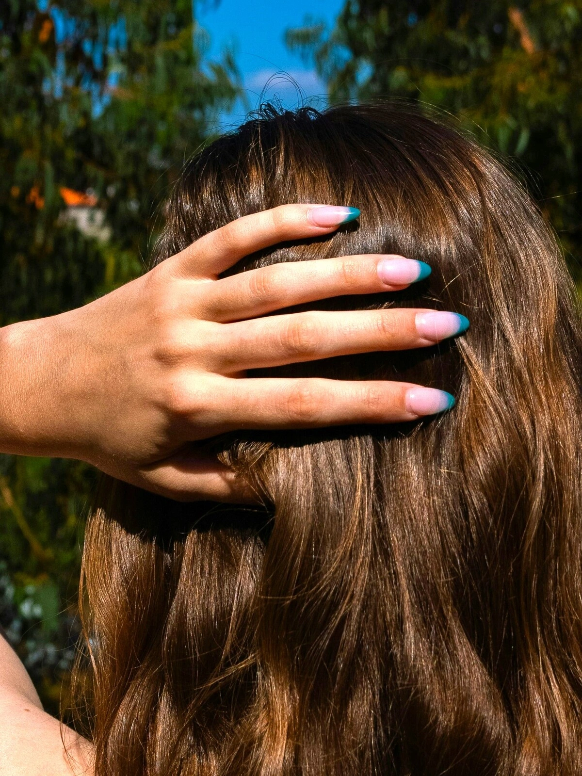 De hand van een vrouw rust tegen haar achterhoofd en toont een French manicure met een lichtroze basis en petrolkleurige tips op amandelvormige nagels