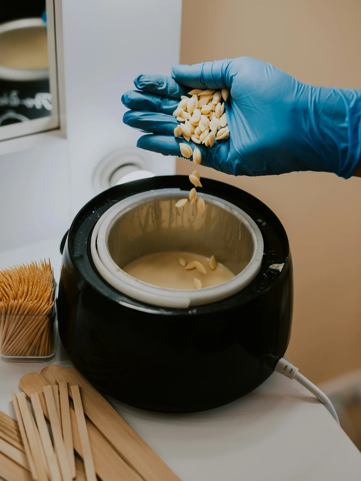 Close-up van de hand van een therapeut met handschoenen die hete waxparels roert in een pot warme hars, klaar voor een klantafspraak