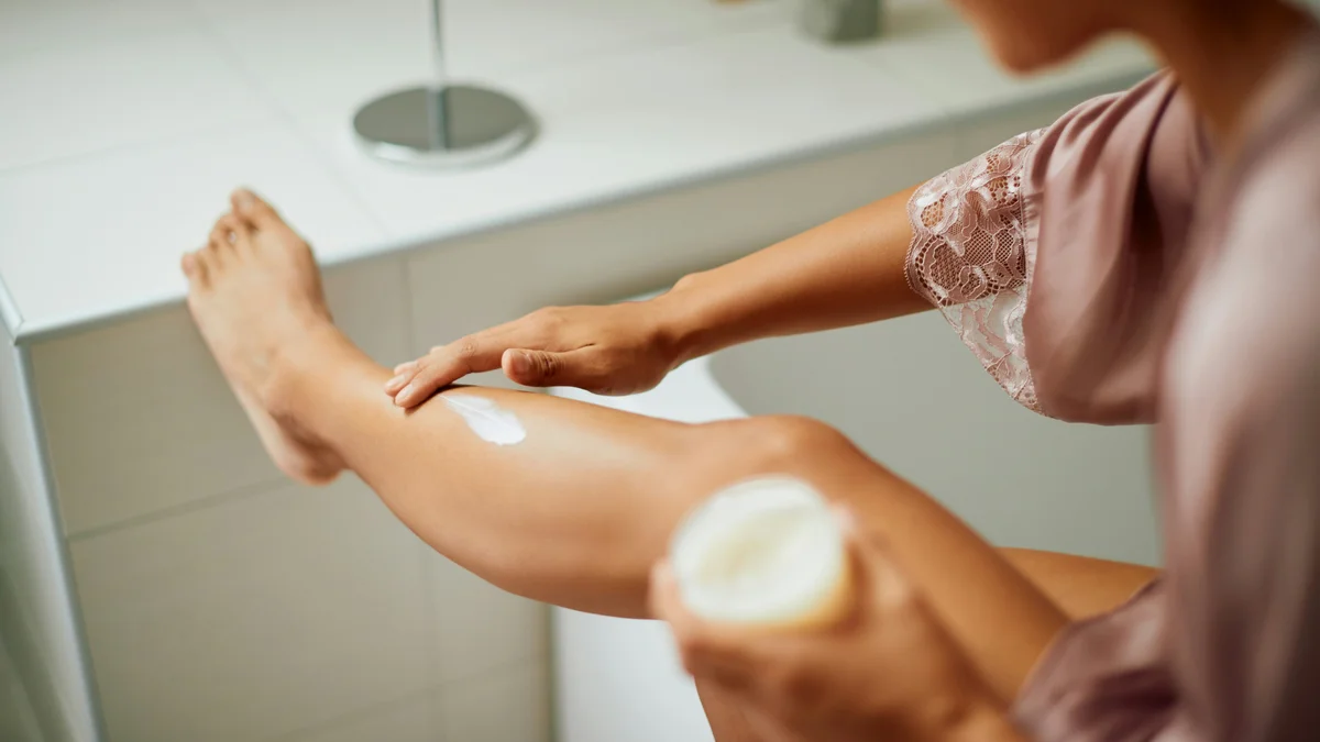 Over-de-schouderopname van een vrouw in een lila zijden ochtendjas die crème op haar been aanbrengt terwijl ze het op het aanrecht in haar badkamer laat rusten