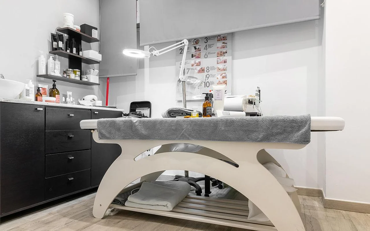 Een compacte behandelcabine bij Atelier Beauty and the Lab, met een crèmekleurige behandeltafel, een verstelbare loeplamp, donkere wandplanken met huidverzorgingsproducten en een genummerd behandelstappen-schema aan de muur.