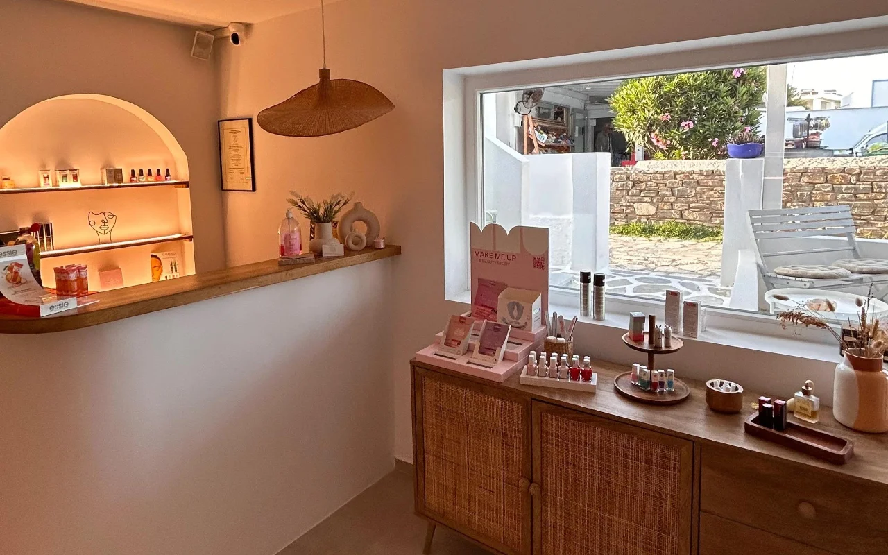 Het interieur van BeautiQ Esthetics and More op Paros, met een zacht verlichte gewelfde nis vol nagellak, een gevlochten rotan hanglamp en een groot raam met uitzicht op een stenen muur en een zonnige tuin.