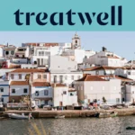 Een witgekalkt vissersdorp aan de Algarve met huizen met terracotta daken die tegen een heuvel omhoogklimmen boven een rustig water, kleine vissersboten afgemeerd langs de kade, palmbomen en een kerktoren onder een strakblauwe lucht.