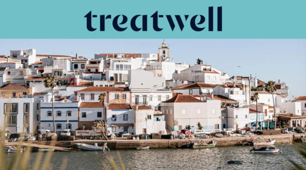 Een witgekalkt vissersdorp aan de Algarve met huizen met terracotta daken die tegen een heuvel omhoogklimmen boven een rustig water, kleine vissersboten afgemeerd langs de kade, palmbomen en een kerktoren onder een strakblauwe lucht.