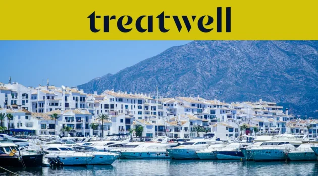 De jachthaven van Puerto Banús in Marbella, omzoomd door witte appartementsgebouwen en afgemeerde jachten met bergen op de achtergrond, onder een geel Treatwell-spandoek.