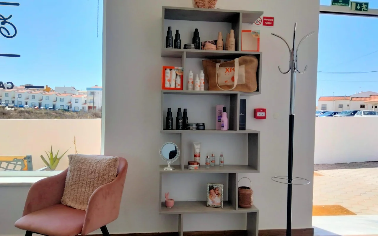 Een salonhoek naast een groot raam, met een getrapte grijze plank vol haarproducten en een ingelijste foto, een oudroze fluwelen fauteuil met kussen, en uitzicht op lage witgekalkte huizen onder een blauwe lucht.