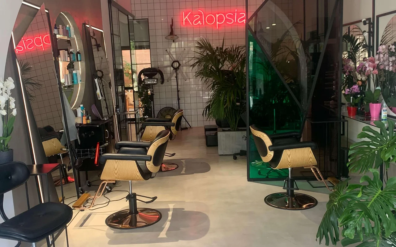 De stylingruimte van salon Kalopsia in Ermoupoli op Syros, met stoelen in licht hout en zwart, gewelfde verlichte spiegels, bladerrijke planten en een roze neonbord met de tekst Kalopsia.