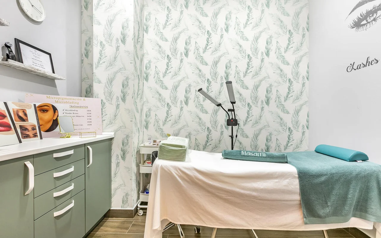 Een behandelkamer bij Masami Belleza Natural in Fuengirola, met een behandeltafel, verenbehang, een vergrootlamp en een prijslijst voor micropigmentatie.