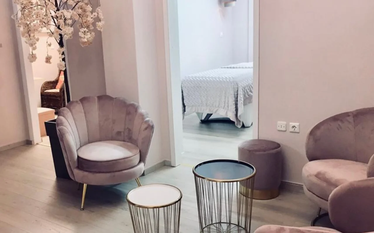 De poederroze wachtruimte van Nomikou Antonia Aesthetic Lab op Syros, met een fluwelen fauteuil met geschulpte rugleuning, dunne gouden bijzettafels, een bloeiende kersentak en een behandelkamer achter de deur.