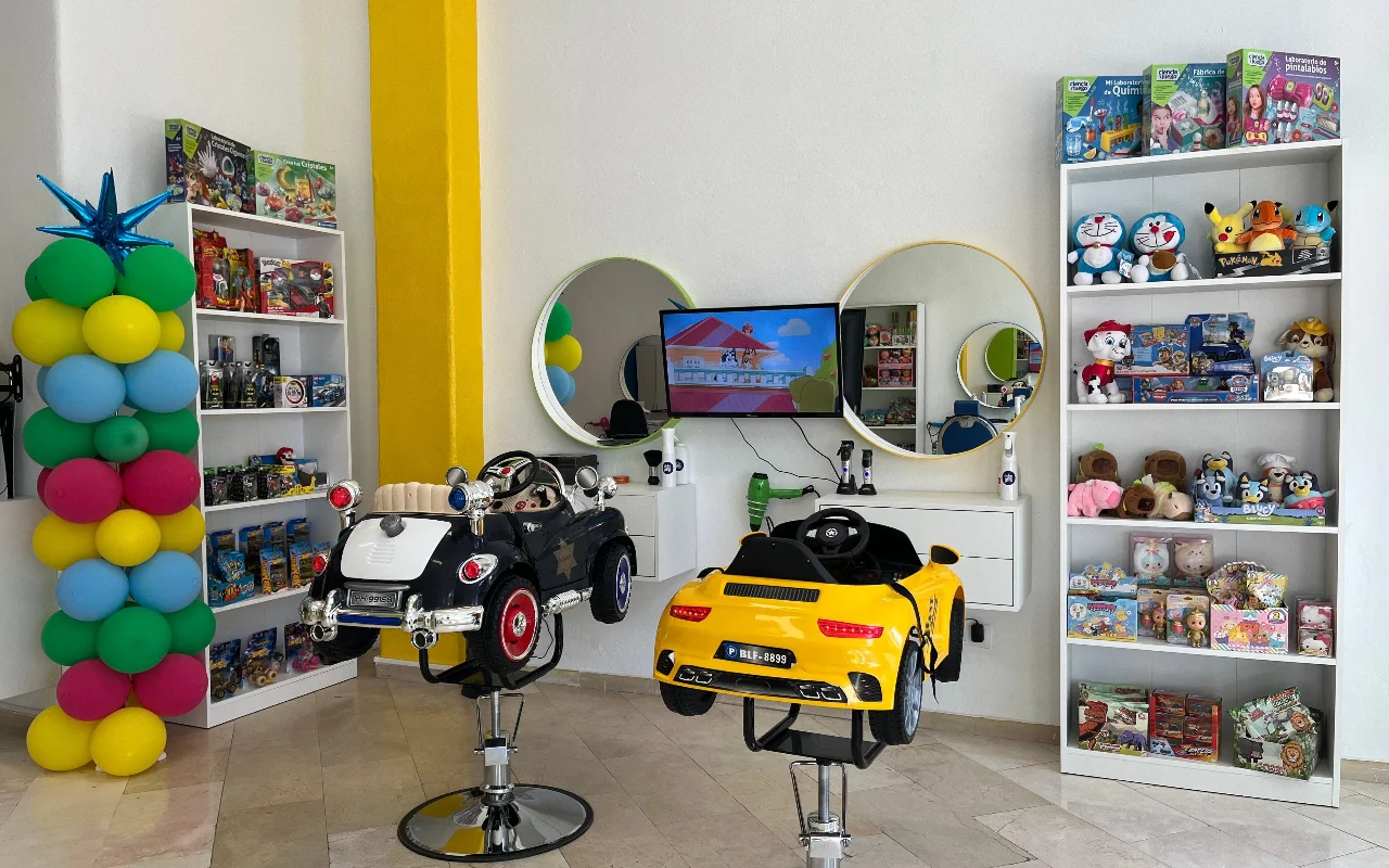 De kinderhoek van Peluplay in Benalmádena, waar de stylingstoelen de vorm van speelgoedauto's hebben, met planken vol tekenfiguren, ronde spiegels en een ballonzuil.