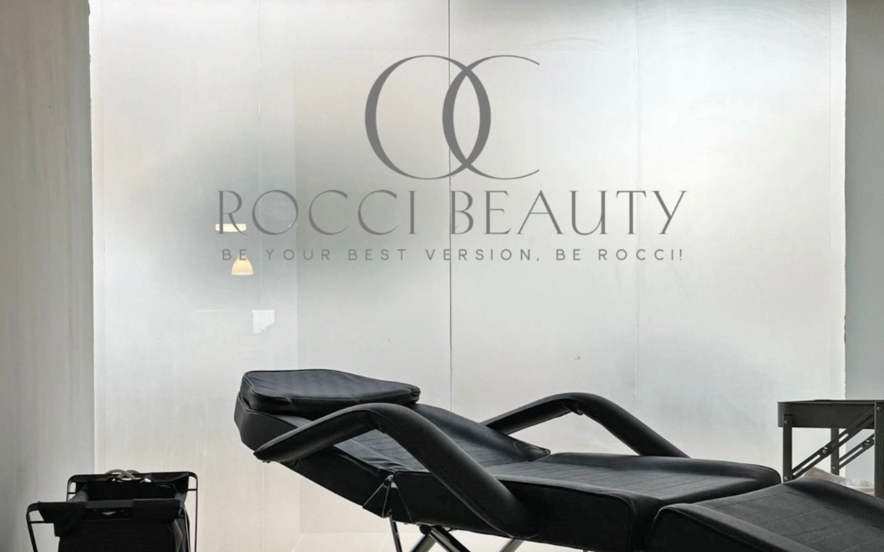 Een behandelruimte met een zwarte verstelbare ligstoel en een klein instrumentenkarretje, tegen een matglazen wand met het logo en de slogan van Rocci Beauty.