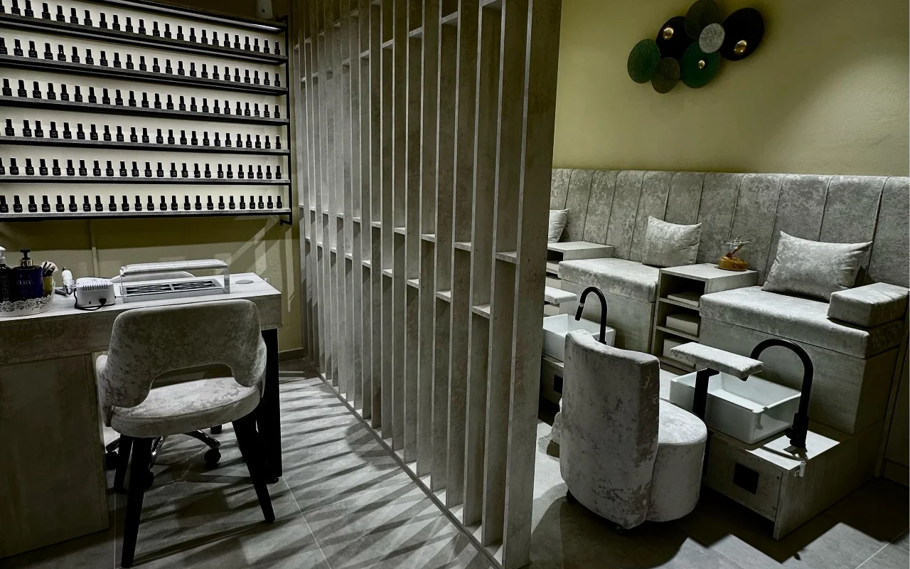 Een behandelkamer bij Sisu Nail Club and Academy in Perissa op Santorini, met een hele wand vol nagellak, grijze fluwelen stoelen voor manicure en pedicure en een opengewerkte betonnen scheidingswand.
