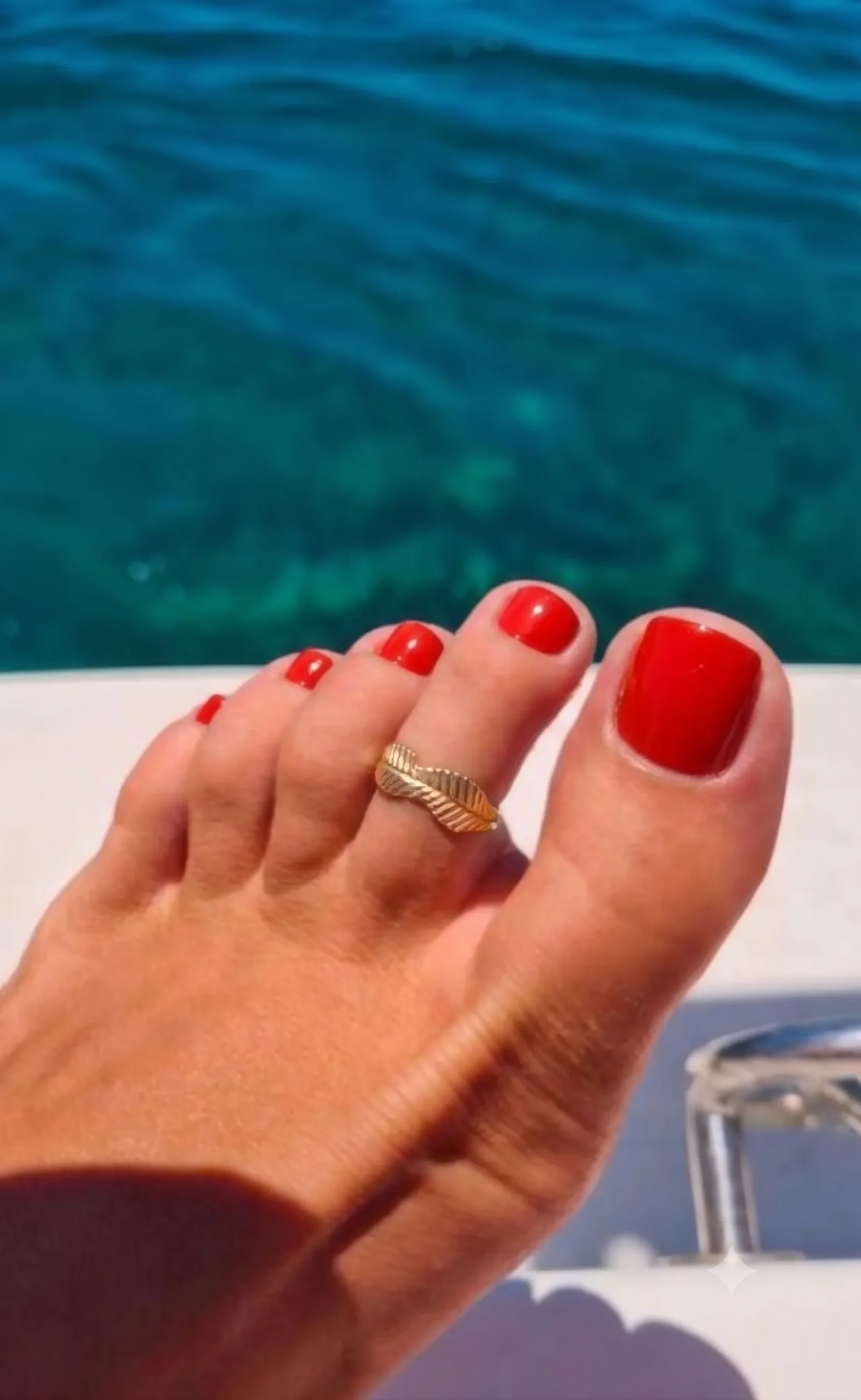 Glanzende tomaatrode pedicure op een gebruinde voet met een gouden teenring, turquoise zee op de achtergrond