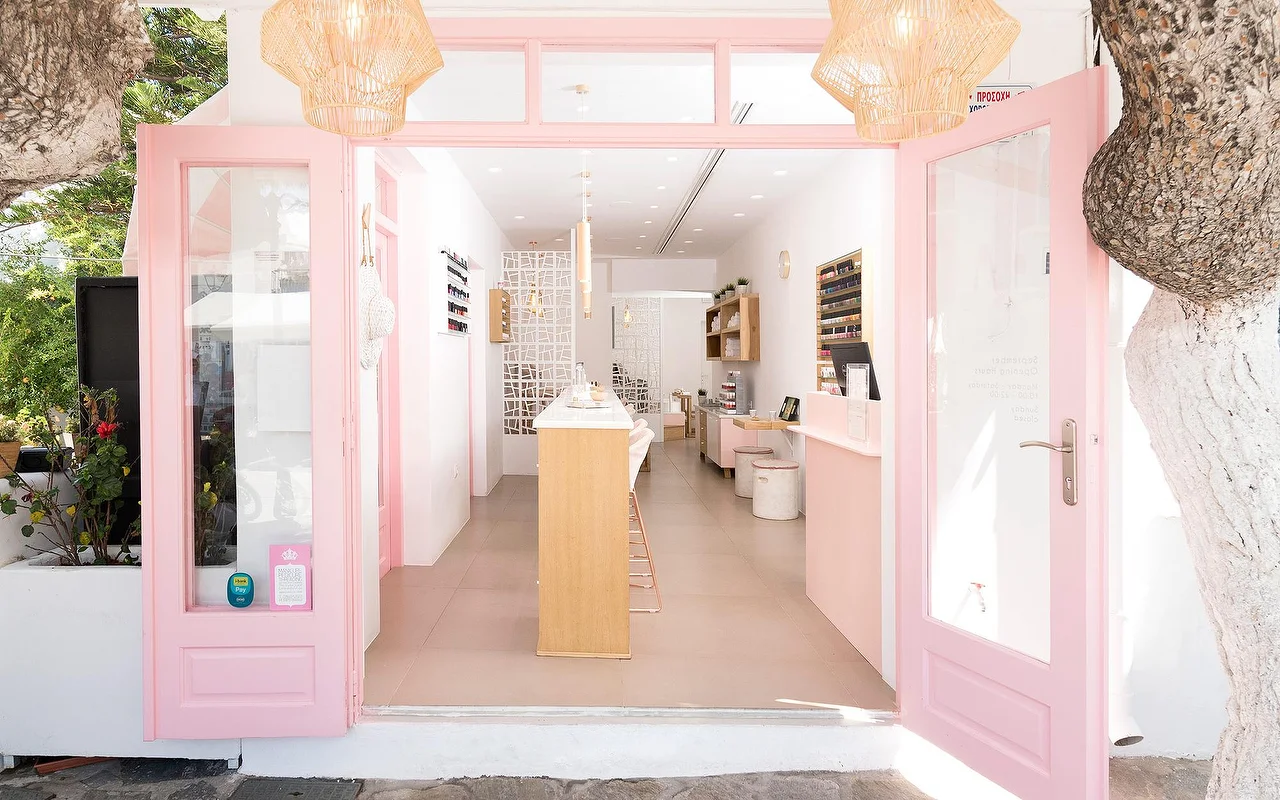 De roze omlijste ingang van Vivian Nail Boutique in Naoussa op Paros, met open deuren, gevlochten hanglampen en een houten toonbank, in een witgekalkt Cycladisch gebouw naast een boom.