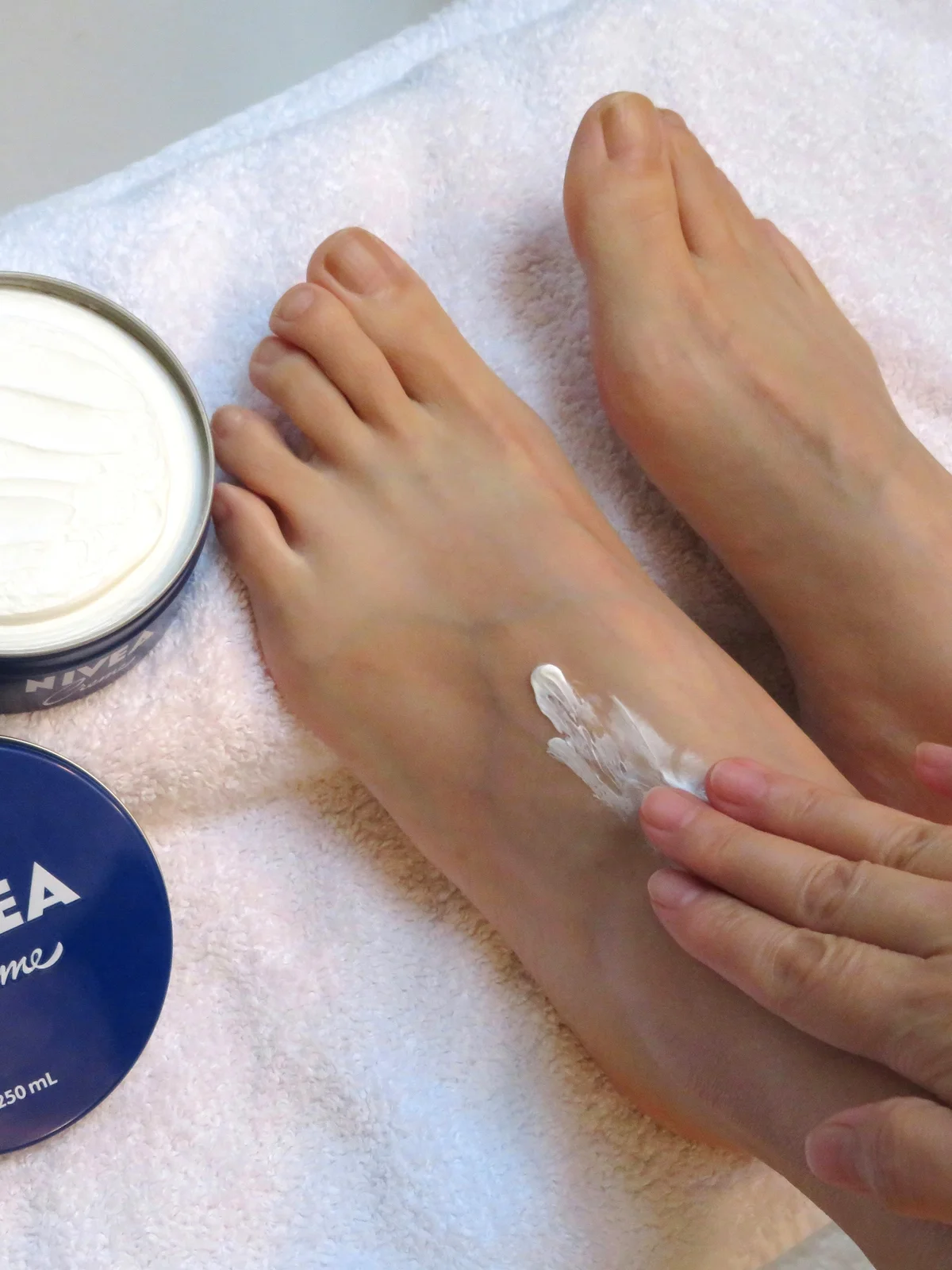 Close-up van de voeten van een vrouw van bovenaf, rustend op een wit laken met een open potje crème ernaast, terwijl ze haar voeten insmeert