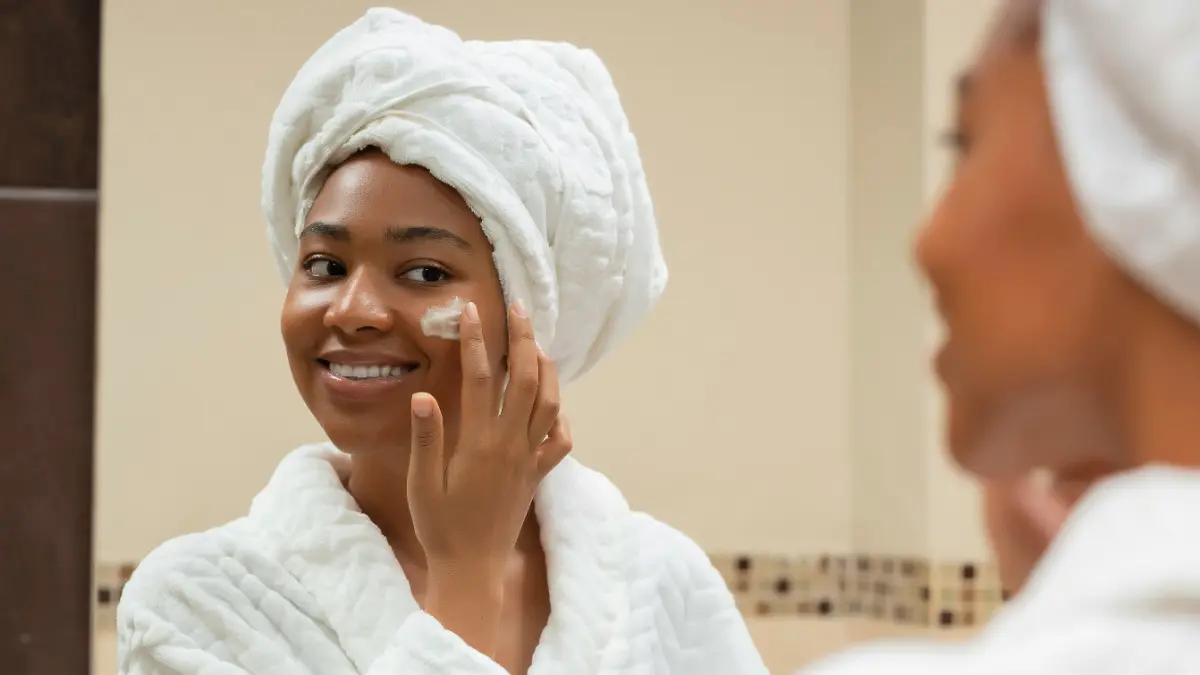 Een vrouw in een witte badjas met een witte handdoek om haar hoofd lacht in de badkamerspiegel terwijl ze SPF op haar gezicht aanbrengt