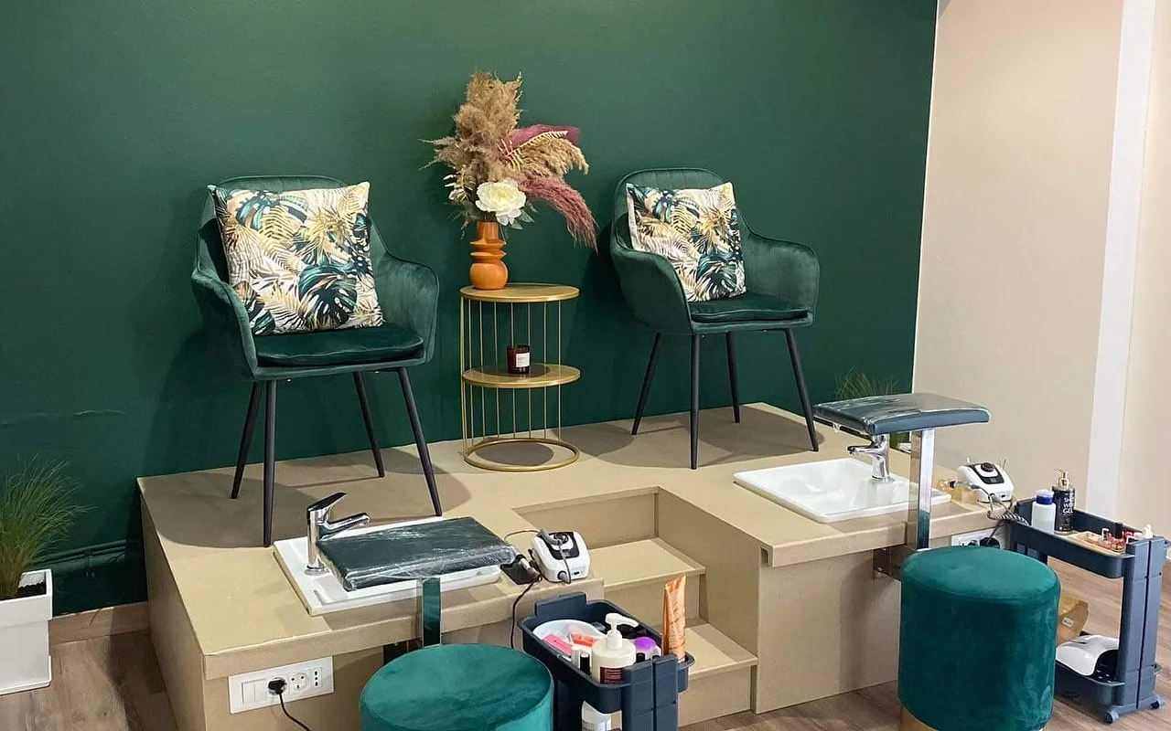 Een pedicure-hoek met een diepgroene muur, twee groene fluwelen kuipstoelen met tropische kussens verhoogd op een beige platform boven pedicurebakken, een gouden bijzettafel met pampasgras in een oranje vaas, en bijpassende groene fluwelen krukken.