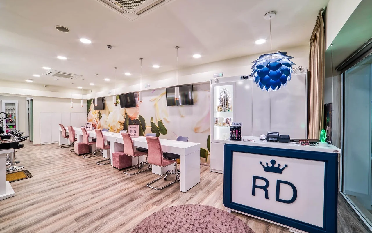 Een helder witte salon met een lange rij manicure-tafels en oudroze stoelen, een zachte florale muurschildering, lichte houten vloer, en een receptiebalie met een 'RD'-kroonlogo.