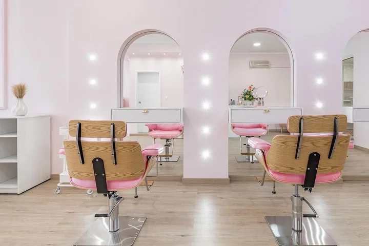 Lichte kapsalon in zachtroze, met houten kappersstoelen met roze bekleding tegenover twee booggewijze, spotverlichte spiegelnissen, witte kasten en vloer met licht houteffect.