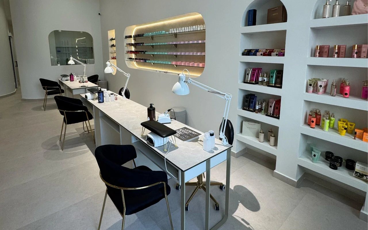 Een witte nagelstudio met manicure-tafels met marmeren blad en witte zwenklampen, zwarte fluwelen stoelen op gouden poten, en boogvormige wandnissen met skincare-producten en een van achter verlichte nagellakrek.