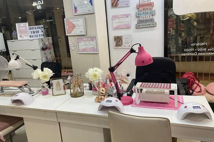 Manicurestation van een nagelsalon met witte tafels, een felroze lamp en LED-lampjes, witte rozen en ingelijste certificaten aan de muur, met de naam Color Nails PT zichtbaar op de ruit naast de openingstijden.