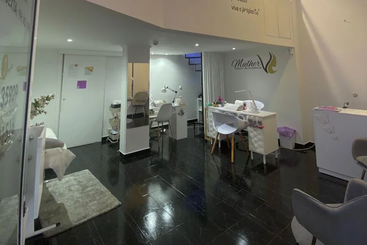 Interieur van een schoonheidsstudio: donkere gepolijste tegelvloer, witte manicuretafel en zachte behandelingszones, het Mulher-logo op de achterwand en een gezichtsbehandelingsstoel zichtbaar achter een glazen wand.