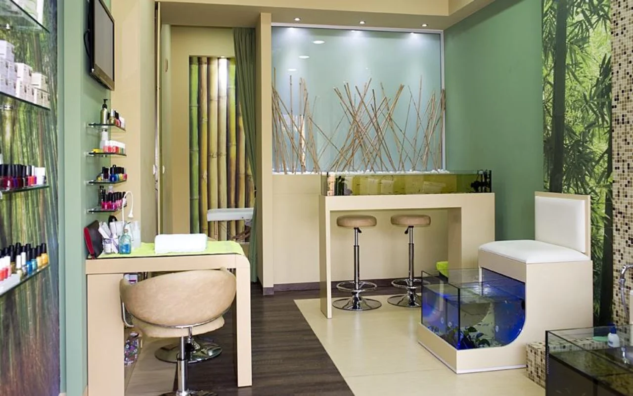 Een compacte salon met bamboe- en bosgroene muurdecoratie, wandplanken met nagellak, een crèmekleurige manicure-tafel en -stoel, twee barkrukken, en een blauw verlicht glazen aquarium voor de fish-spa-pedicure.