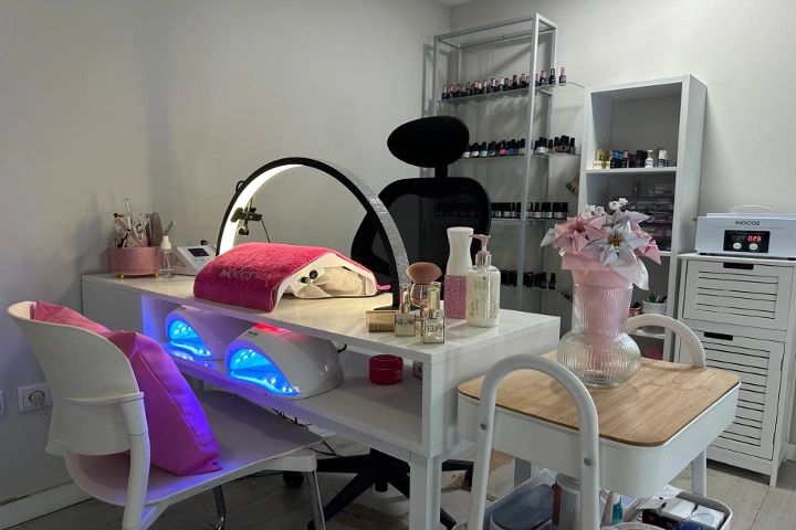 Enkele nagelplek met een witte tafel, een ringlamp en twee blauw oplichtende LED-lampen, een zwarte stoel en planken vol kleurige nagellak, opgefleurd door een roze vaas met bloemen.