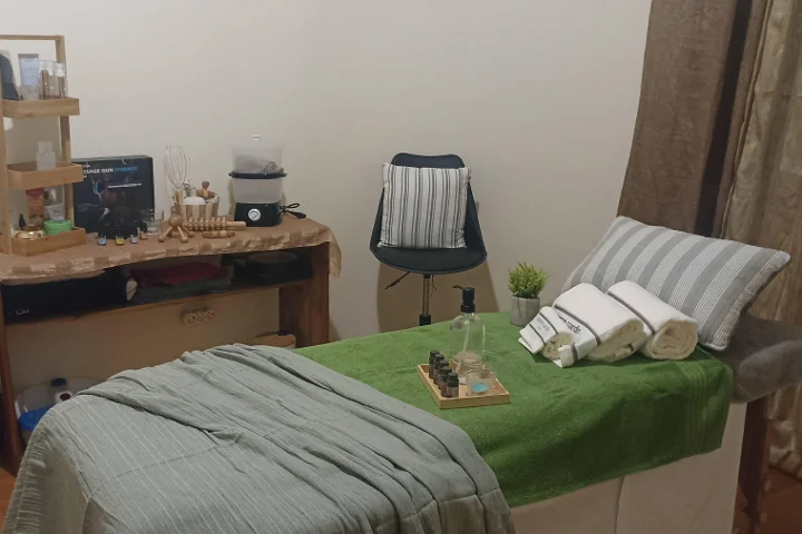 Gezellige massagekamer met een tafel bedekt met een groene handdoek en opgerolde witte handdoeken, een schaal etherische oliën, houten massagetools op een rustiek bijzettafeltje, en een kruk naast een gordijnenhoekje.