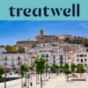 Zonnig uitzicht over Ibiza-Stad, met witgekalkte gebouwen die oplopen naar de kathedraal en de oude vestingmuren van Dalt Vila op de heuvel. Een door bomen omzoomde havenpromenade loopt door de voorgrond onder een strakblauwe hemel.