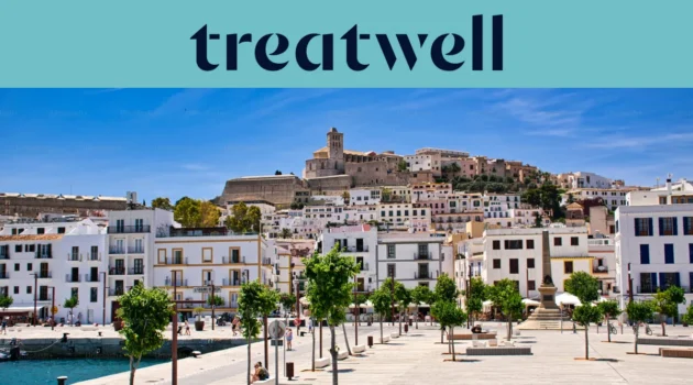 Zonnig uitzicht over Ibiza-Stad, met witgekalkte gebouwen die oplopen naar de kathedraal en de oude vestingmuren van Dalt Vila op de heuvel. Een door bomen omzoomde havenpromenade loopt door de voorgrond onder een strakblauwe hemel.