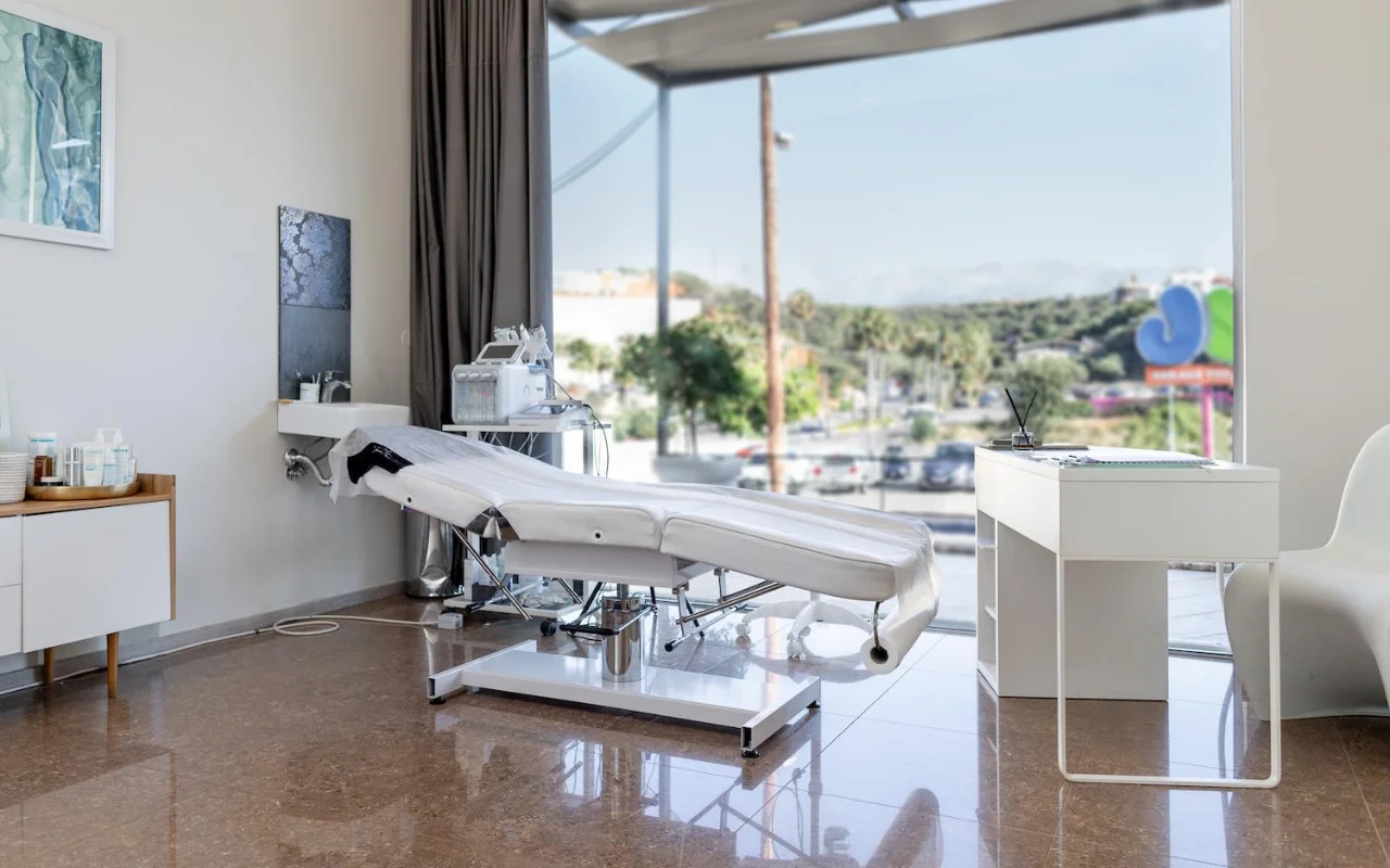 Een schone witte behandelkamer met een verstelbare beauty-bank, skincare-apparatuur op een trolley, ingelijste kunstwerken en een groot raam met uitzicht op bomen, een weg en heuvels in de verte.