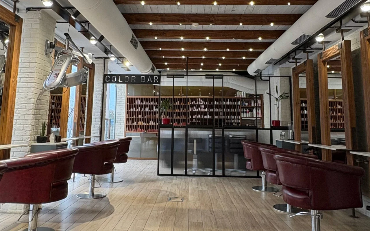 Een kapsalon met blootliggende houten dakbalken en witgekalkte baksteen, bordeauxrode leren stylingstoelen en een kleurenkamer met glazen wanden vol productplanken achter een 'Color Bar'-bord.
