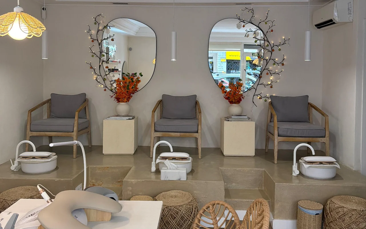 Een lichte, moderne nagelsalon met twee pedicurestoelen op een verhoogd betonnen platform, elk met een wit voetbad en een grijze gestoffeerde fauteuil. Twee organisch gevormde spiegels, versierd met takken met lichtjes en oranje bloemen, hangen erboven, met een manicurestation op de voorgrond.