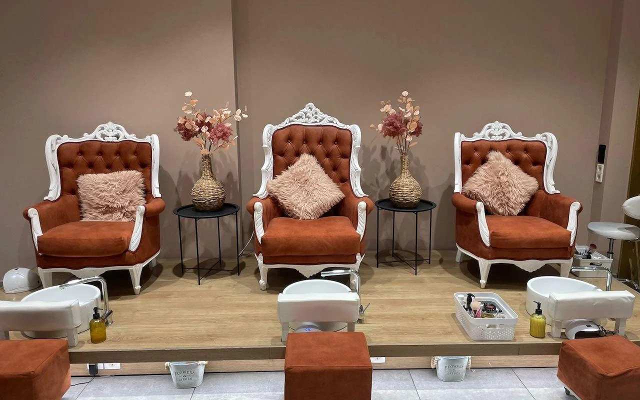 Drie sierlijke wit omlijste stoelen in verbrand oranje fluweel, verhoogd op een houten platform boven witte pedicurebakken, met gedroogde bloemen en zachtroze kussens tussen de zetels.
