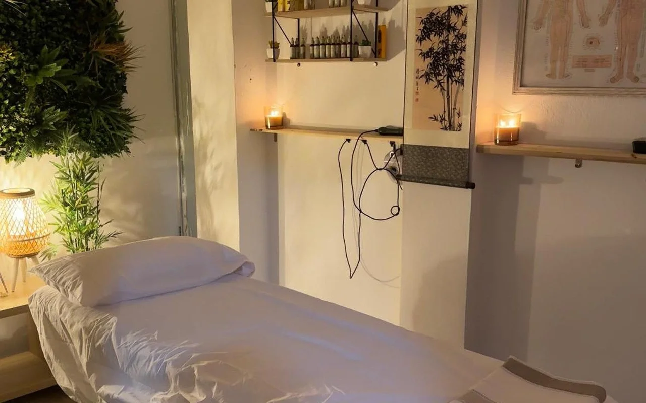 Een sfeervol verlichte behandelkamer met een massagebed opgemaakt in wit linnen. Aangestoken kaarsen staan op houten wandplanken naast een bamboe-inktrol en een ingelijste acupunctuurkaart, met een groene plant en een gloeiende gevlochten lamp in de hoek.