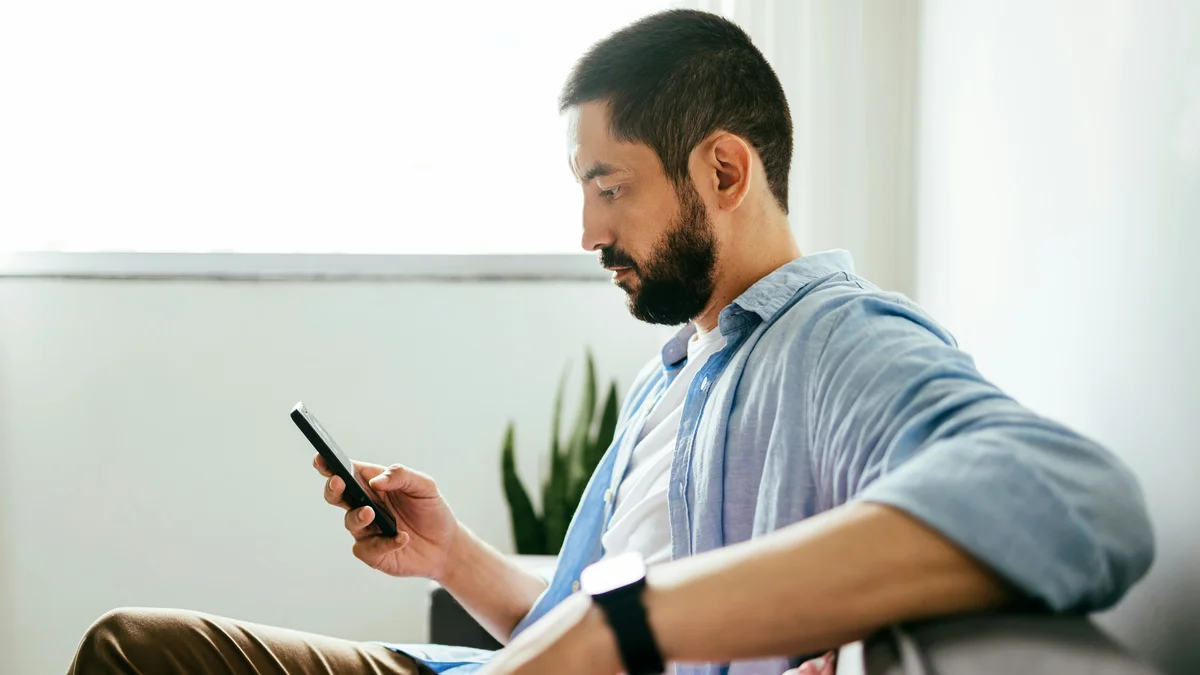 Zijaanzicht van een man die op zijn bank zit, met één arm op de rugleuning, en met zijn andere hand op zijn telefoon een behandeling boekt